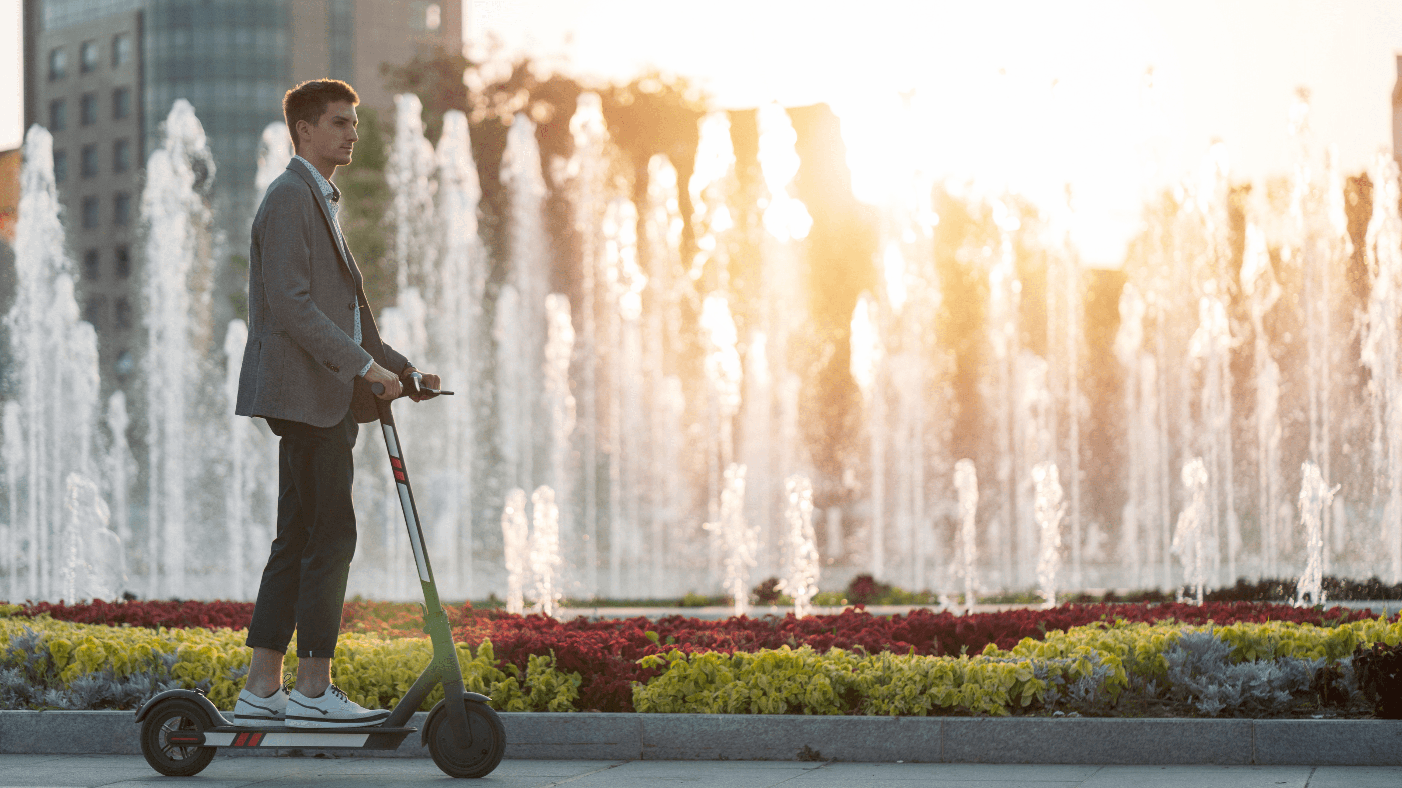 Junger Mann auf E-Scooter vor einem Brunnen in einer Großstadt mit Hochhaus im Hintergrund.