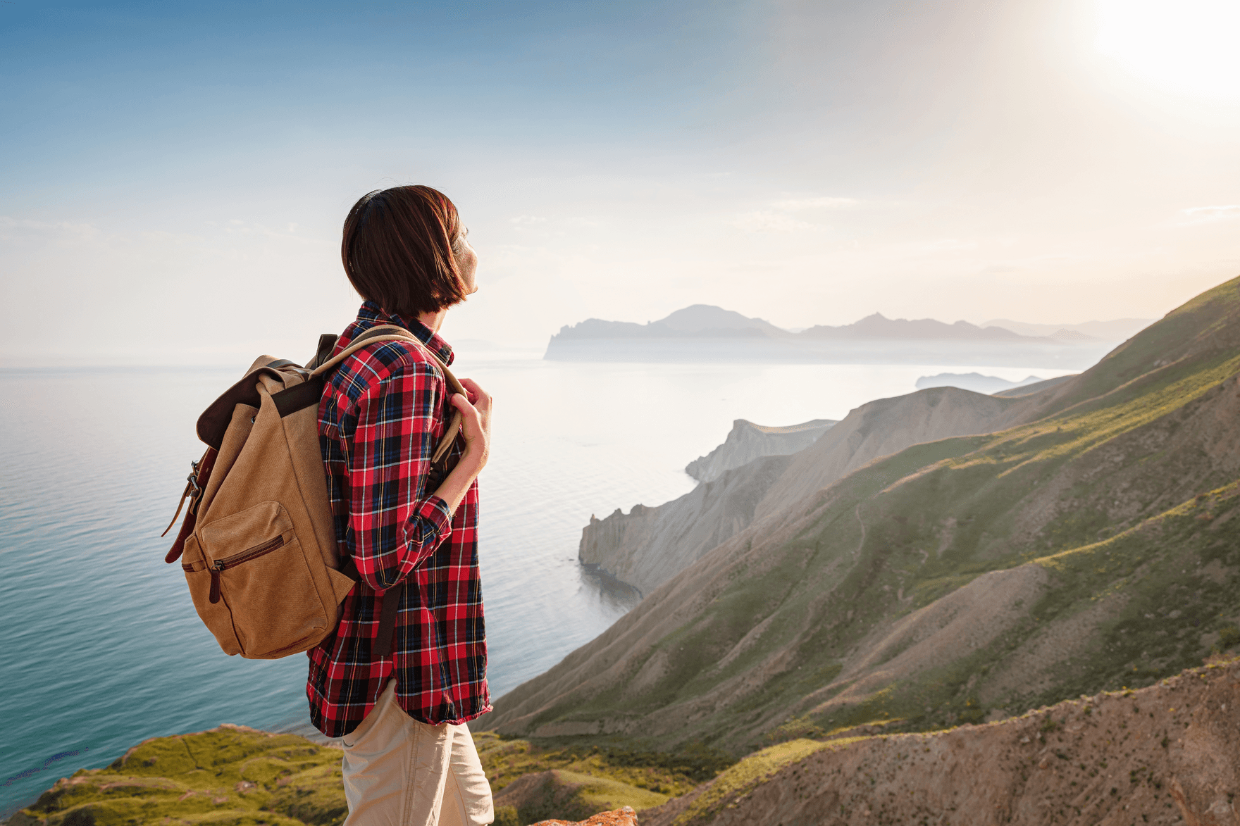 Eine Frau mit Rucksack steht auf einem Küstenwanderweg und blickt in die Ferne über Meer und Berge.